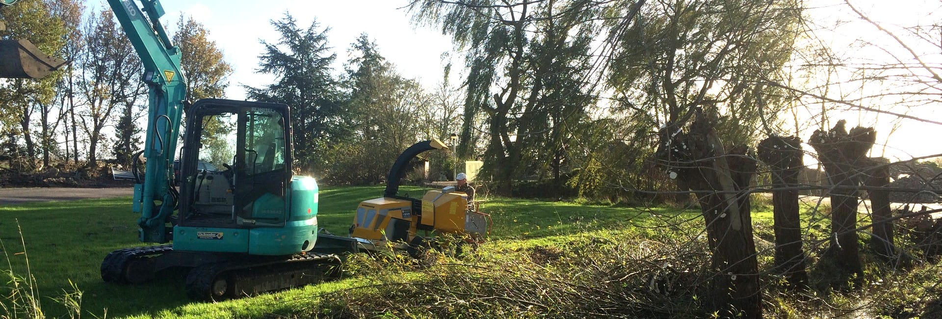 Lichtblauwe minikraan en gele houtversnipperaar die snoeihout ruimen op een grasveld
