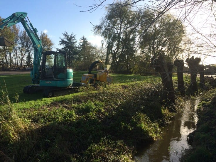 Lichtblauwe minikraan en gele houtversnipperaar langs een beek met treurwilgen