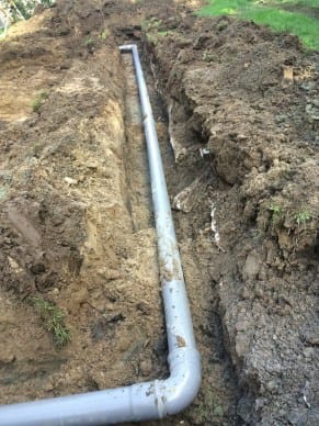 Grijze rioleringsbuis netjes gelegd in een diep uitgegraven aardesleuf in een tuin