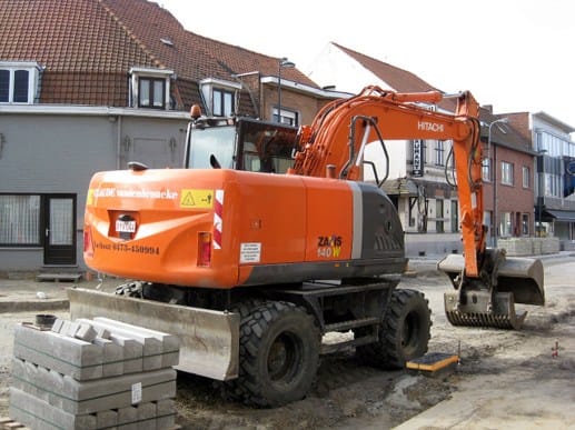 Oranje Hitachi rupskraan aan het werk in een dorpsstraat met betonkeien