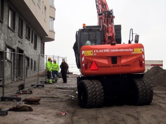 Oranje mobiele Claude-kraan op een stedelijke bouwwerf met arbeiders in fluogeel